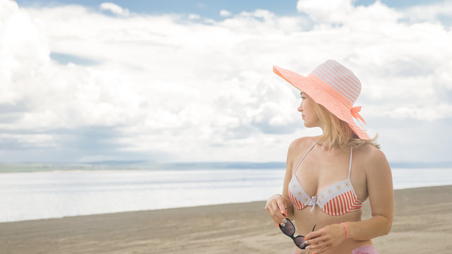 Attractive woman in a hat stands on the beach against the blue sea. She has sunglasses and a panama on her head. On her body is a swimsuit and a silk cape. In the background are very beautiful deep