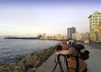 Photographer with camera during sunset with Atlantic Ocean, residential building and Morro Castle in background - Malecon, Havana, Cuba