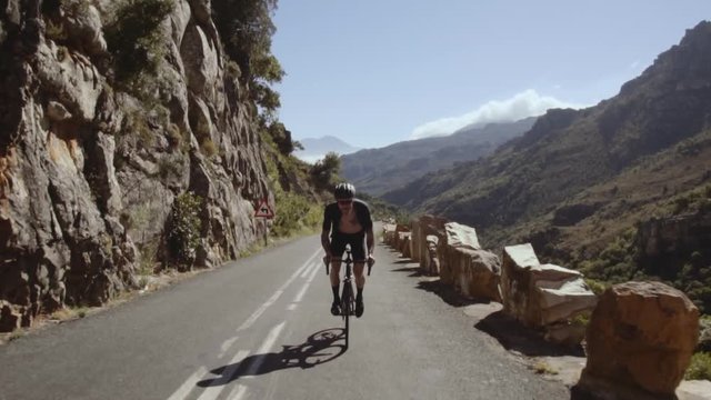 Tracking shot of a male cyclist doing uphill ride. Sportsman climbing up the curvy hilly road on a bicycle. 
