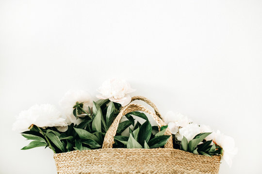 Fototapeta Straw bag with white peony flowers on white background. Flat lay, top view summer floral concept.