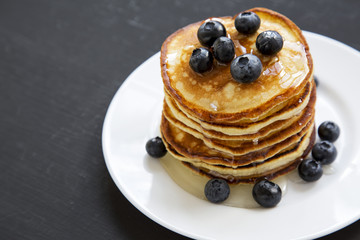 Pancakes with blueberries and honey on a white round plate over black wooden background, side view. Close-up. Copy space.