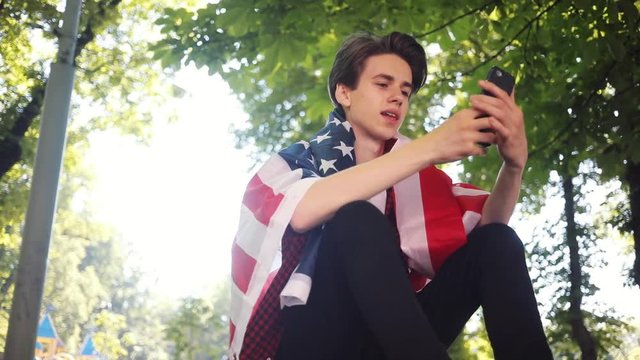 Close up boy with American flag using mobile phone smile sitting on skate in park guy business fashion hand child man nature internet social smartphone face lifestyle smart teenager young slow motion