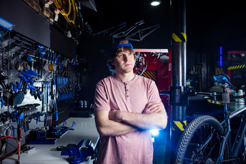 Small business and bicycle transport service. Portrait of a young man in a cap posing against the backdrop of a bicycle workshop and a tool for setting up and repairing
