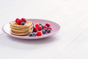 Delicious homemade pancakes with fresh berries in a violet plate on a white background. A tasty and healthy breakfast of pancakes with raspberries and blueberries.