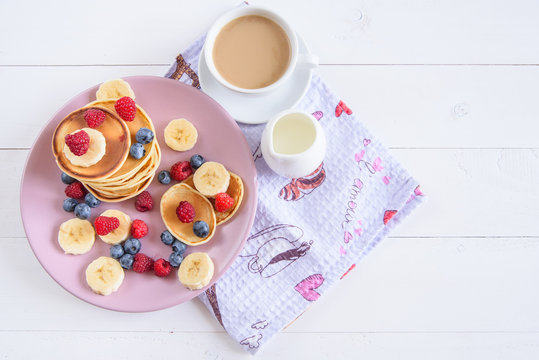 Delicious homemade pancakes with fresh berries in a violet plate on a white background. A tasty and healthy breakfast of pancakes with raspberries, blueberries and bananas. Aromatic coffee with milk.