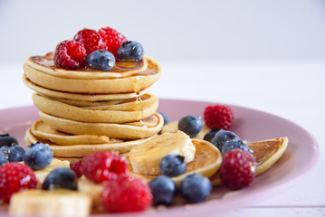 Delicious homemade pancakes with fresh berries in a violet plate on a white background. A tasty and healthy breakfast of pancakes with raspberries, blueberries and honey.