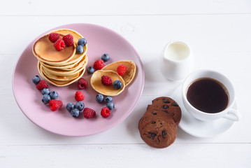 Pancakes with fresh berries in a violet plate on a white background. A tasty and healthy breakfast of pancakes with raspberries, blueberries and fresh milk. Fragrant coffee with cookies.