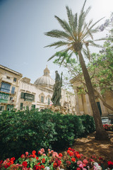 Fototapeta na wymiar Beautiful view from square with green bushes. flowers and palm of satue of saint and cathedral dome, church in Valetta, Malta