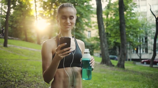 Happy young woman use smartphone and headphone drink water in park sun outdoor health hand girl tree sport fitness summer nature internet technology runner exercise phone workout mobile cellphone
