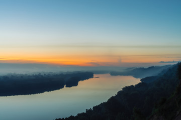 View from high shore on river. Riverbank with forest under thick fog. Dawn reflected in water. Yellow stripe in picturesque predawn sky. Colorful calm morning atmospheric landscape of majestic nature.