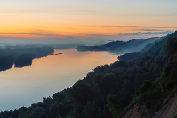 View from high shore on broad river. Riverbank with forest under thick fog. Dawn reflected in water. Yellow glow in picturesque predawn sky. Colorful morning atmospheric landscape of majestic nature.