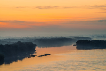 Morning mystic haze above broad valley of river. Gold glow from dawn in sky. Riverbank with forest under fog. Sunlight reflected in water at sunrise. Colorful atmospheric landscape of majestic nature.