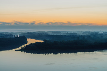 Early blue sky reflected in river water. Riverbank with forest under predawn sky. Yellow stripe in picturesque sky. Fog hid trees on island. Mystical morning atmospheric landscape of majestic nature.