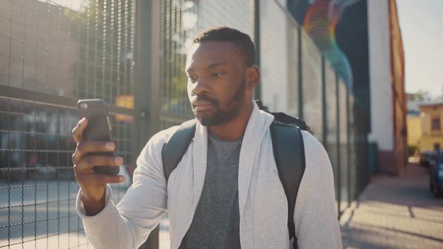 Nice modern guy in casual clothes walking in the street, using smartphone, touching screen. Focused African young man in the street of beautiful city. Outdoors.