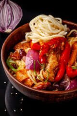 Japanese, Chinese udon noodles with chicken and vegetables, Bulgarian pepper, red onion, white and black sesame. dish in a ceramic dish made of red clay on a black background.