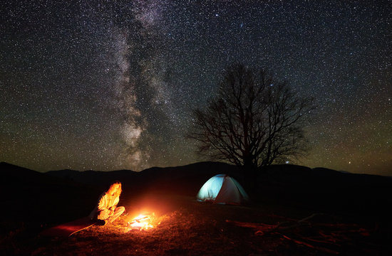 Camping night in mountains. Young female hiker having a rest near burning campfire under incredible starry sky and Milky way with glowing tent and silhouette of big tree in background