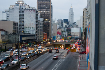 A crowded avenue with car with lights on and the metropolis with a white tall tower in the background.