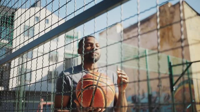 Good-looking young African man playing with basketball in sports ground on background of houses with colored graffiti. Likable guy on holidays. Summertime. Daytime.