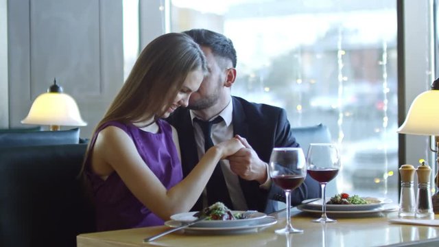 PAN of bearded man in suit sitting at table in restaurant and flirting with girlfriend on date: he holding her hand and whispering compliments in her ear