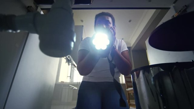 Shot from inside of kitchen sink cabinet of black woman with flashlight inspecting pipes and talking on mobile phone with plumber giving her instructions