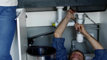 Tilt up of black female homeowner with mobile phone talking to male plumber with wrench lying under kitchen sink and working with pipes