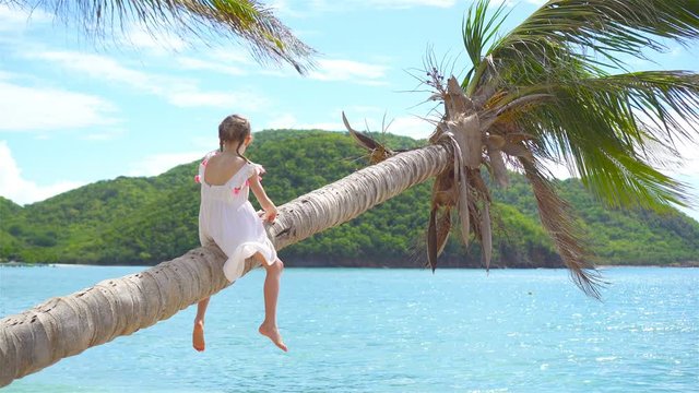 Adorable little girl at tropical beach sitting on palm tree during summer vacation