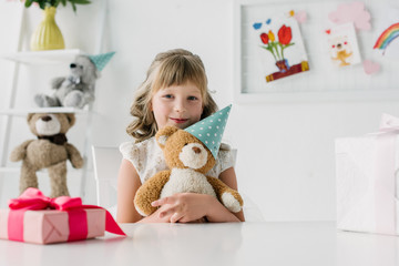cute birthday child holding teddy bear in cone at table with gift box