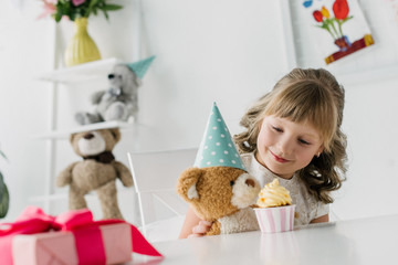 cute birthday kid feeding teddy bear in cone by cupcake at table