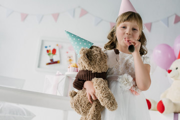 happy birthday child in cone blowing in party horn and holding teddy bear