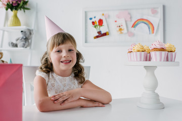 Fototapeta na wymiar cute birthday kid in cone looking at camera and sitting at table with cupcakes