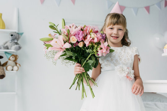 smiling birthday kid in cone looking at camera and holding bouquet of lilies