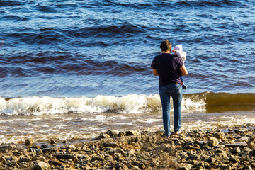 A man with a small child in his arms is standing on the stones on the river bank and looking at the waves..