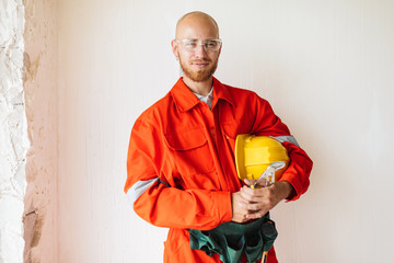 Young foreman in orange work clothes and protective eyewear holding hardhat and tools in hand happily looking in camera over white background