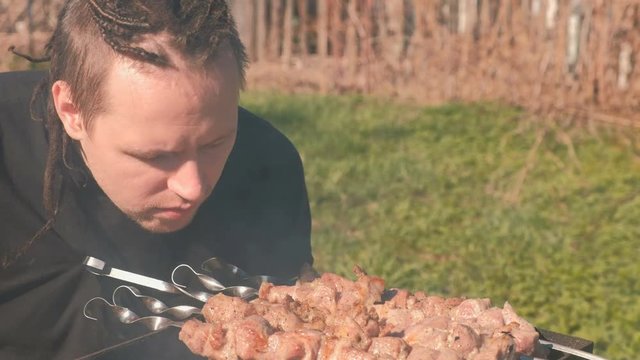 Young men with dreads on hairs cooks shashlik meat on top of charcoal grill on backyard.