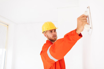 Young foreman in orange work clothes and yellow hardhat thoughtfully using putty knife at work...