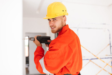 Young foreman in orange work clothes and yellow hardhat dreamily looking in camera using drill at work