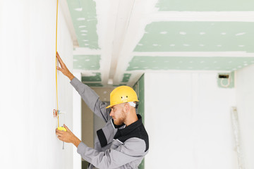Young builder in work clothes and hardhat using measuring tape spending time at work