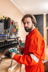 Builder in work clothes thoughtfully looking in camera holding hammer in workshop