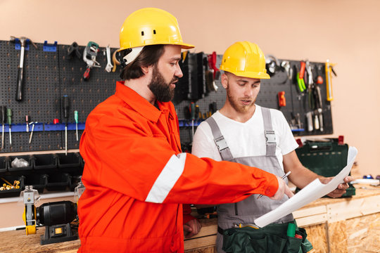 Two builders in work clothes and yellow hardhats thoughtfully looking on plan with tools on background in workshop