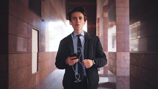 Good-looking young Caucasian man with comely haircut walking near building in big city. Attractive Ukrainian guy with headphones listening to favorite music, enjoying. Lunch break.