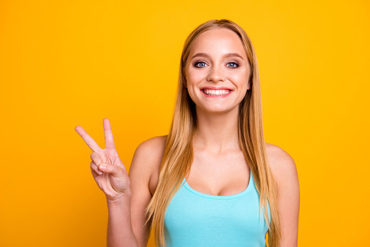 Portrait of young woman with blond hair, big blue eyes and toothy beaming smile. Positive girl demonstrating two fingers v-sign isolated on yellow background with copy space