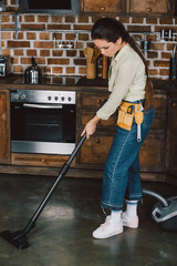 Fototapeta na wymiar attractive young woman with toolbelt using vacuum cleaner at kitchen