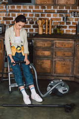 Fototapeta na wymiar sad young repairwoman looking at broken vacuum cleaner while sitting on stepladder at kitchen