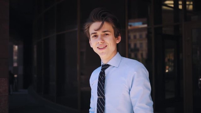 Cute Caucasian guy with smart watch looking at screen of smartphone, using device. Young attractive man with comely haircut looking at camera, smiling. Outdoors.