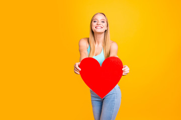 Happy and cute young blond woman expresses her feelings holding red heart in her hands isolated on yellow background