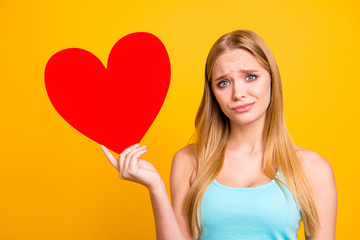 Portrait of sad and serious girl holding big paper carton heart figure. Young blond woman holding red heart in her hands isolated on yellow background