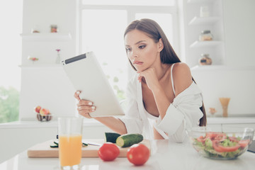 Obraz na płótnie Canvas Sexy woman on white lingerie and long shirt with naked shoulder sit in thought, look at the tablet and biting lower lip. Kitchen with cutting board, ingredients for salad and juice on the table