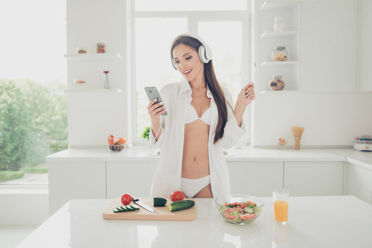 Charming and beautiful young woman in white lingerie and shirt, listening music through headphones, looking on the phone and holding fork on the hand cooking vegetable salad for breakfast
