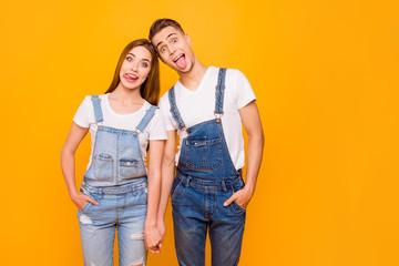 Naklejka premium Portrait of funny fool playful lovely adorable young cute couple holding hands, showing tongues out, standing straight over yellow background