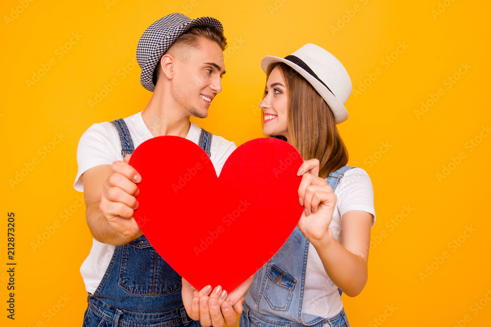 Wall mural Young happy beautiful students couple in love wearing hats holding red paper heart, isolated